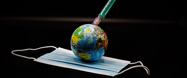 Photo of ball, representing the Earth, on top of a COVID mask with vaccination syringe inserted in the Earth.